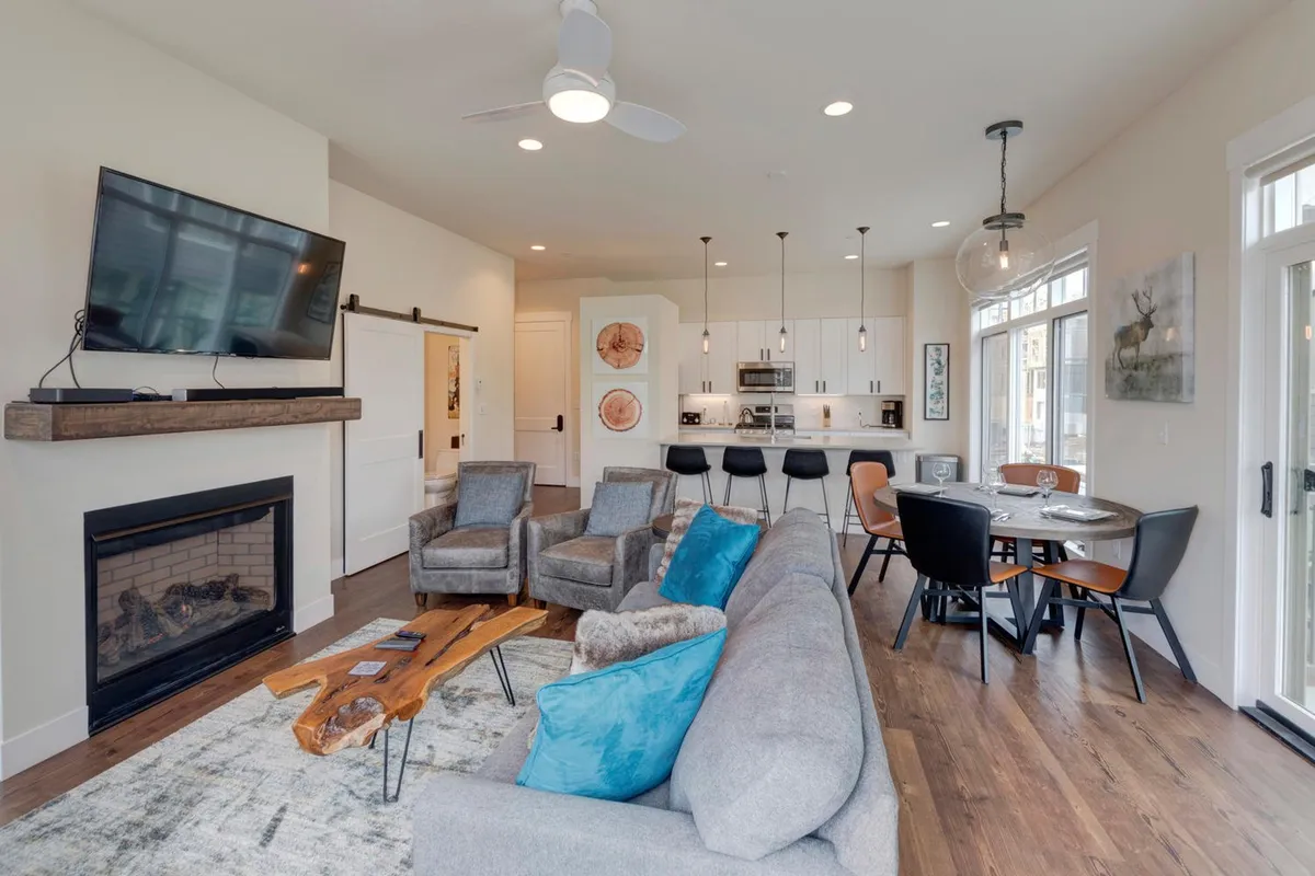 The image showcases a welcoming, contemporary open living area with a glowing fireplace as the centerpiece, surrounded by a plush sofa, a stylish kitchen island, and an inviting dining table. The scene exudes warmth and comfort, perfect for gathering, relaxing, or sharing memorable moments indoors.