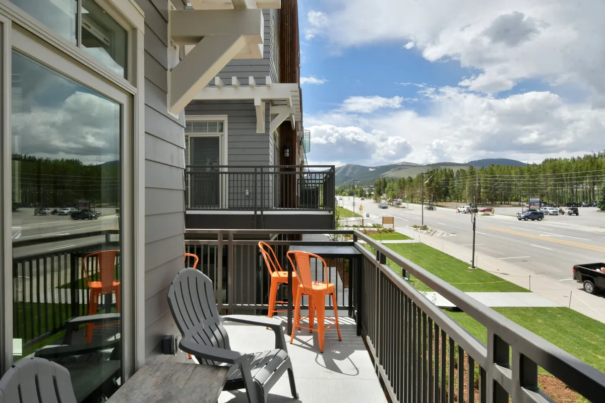 This image shows a comfortable balcony seating area overlooking a stunning mountain landscape, with ample space for relaxing outdoors. The scene captures a moment where someone could enjoy morning coffee or relax with friends while taking in the sweeping views. The visible street below hints at easy access to the vibrant local scene.