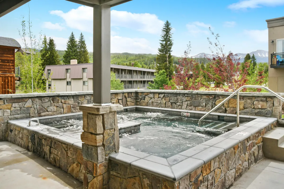 This image features an expansive outdoor hot tub set against a scenic backdrop of mountains and trees. Guests are seen relaxing in the hot tub, enjoying serene views and a tranquil atmosphere, with social interactions among friends hinting at a communal, resort-like amenity.