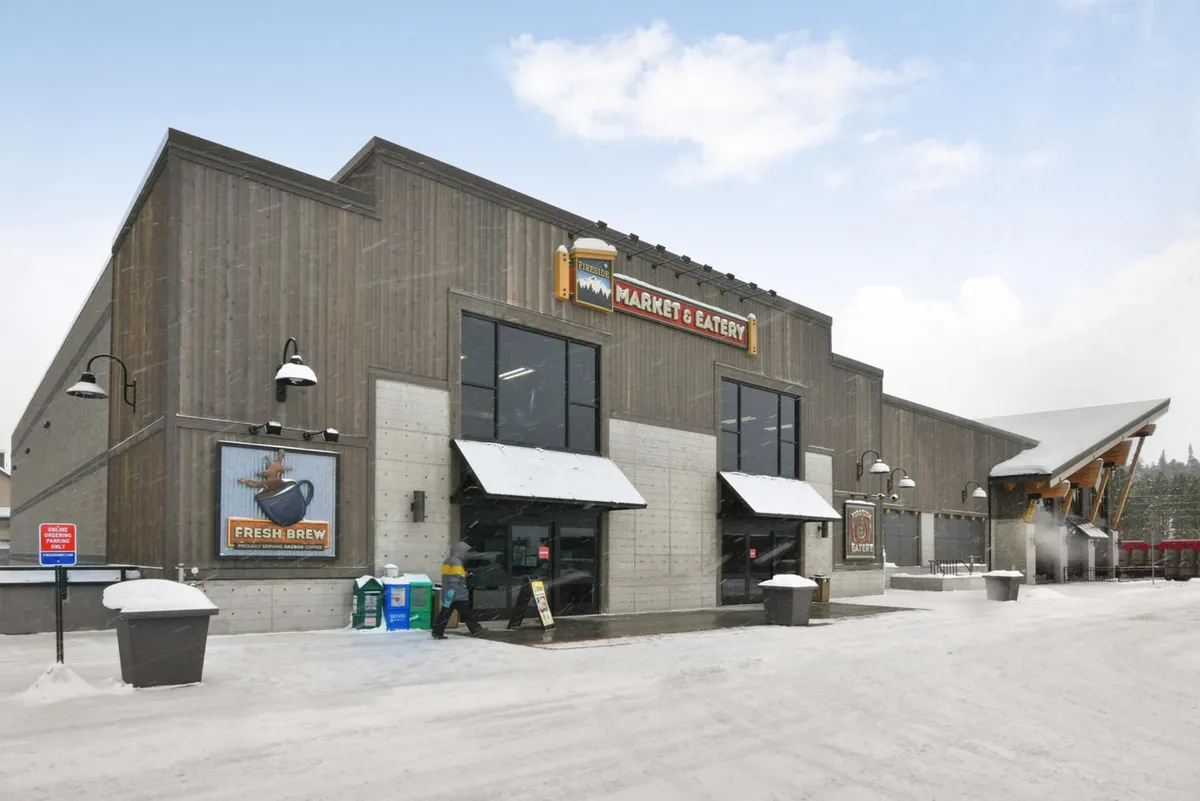 The image shows the welcoming entrance to the Fireside Market, with a visible market sign, snow-covered ground, and an inviting outdoor seating area. The mood is warm and communal, evoking a sense of convenience and neighborhood spirit.