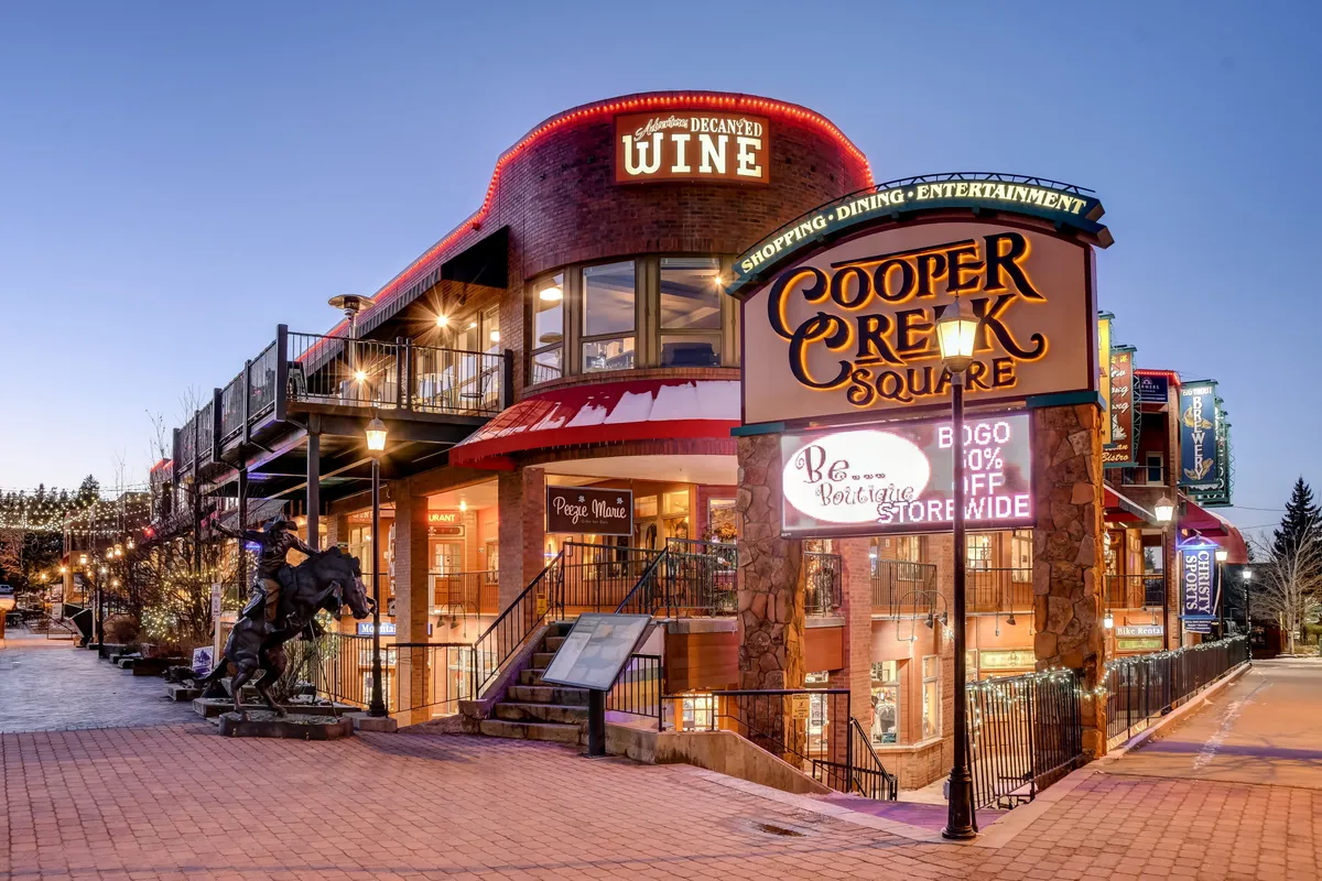 A lively exterior shot of Cooper Creek Square, featuring the prominent sign, outdoor dining areas, boutique shops, and people socializing. The setting is energetic and vibrant, with touches that suggest live music and community gatherings, all set against the backdrop of a mountain town.