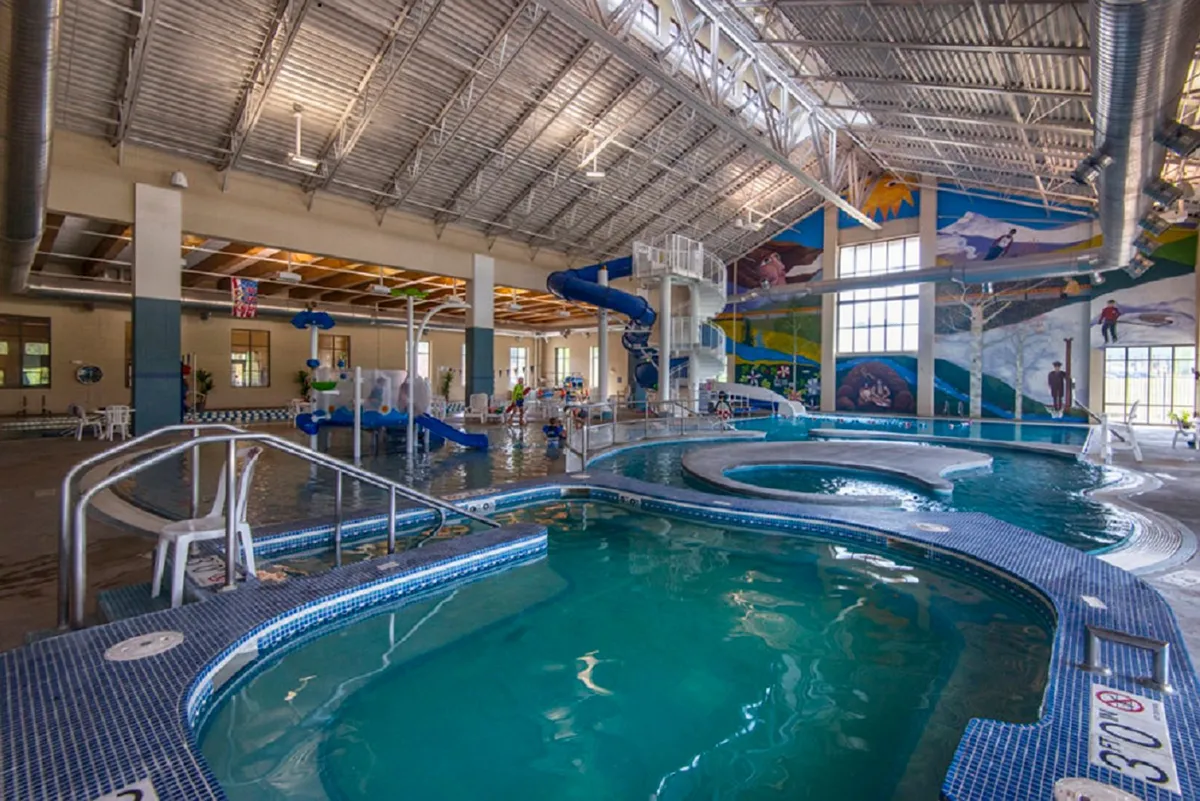 An energetic indoor pool and play area featuring a large water slide, colorful murals, and children playing. The scene is joyful, depicting family fun and the recreational facilities mentioned, such as those at the Grand Park Rec Center.