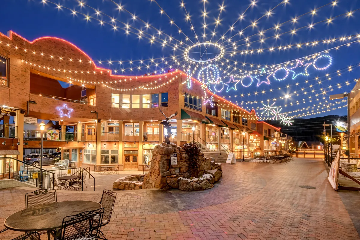 A lively, festive outdoor plaza lit by string lights and decorated with stars, featuring a stone fountain and numerous seating areas. People are dining, shopping at market stalls, and celebrating, capturing the joy and energy of community events and downtown life.