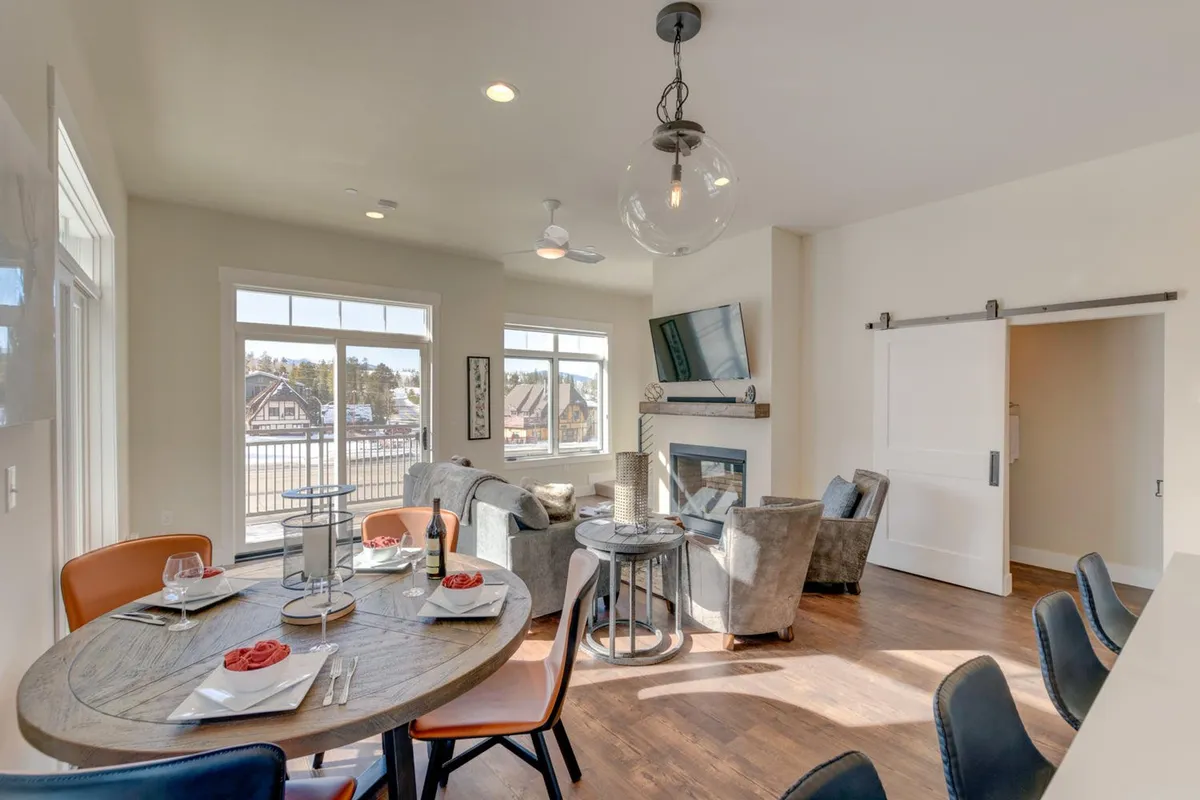 A bright and welcoming living space filled with natural light from expansive windows. The focal points include a modern gas fireplace, a spacious dining table, and comfortable seating arranged to encourage conversation or relaxation. The view through the windows hints at outdoor access, possibly the wrap-around balcony mentioned in the narrative.