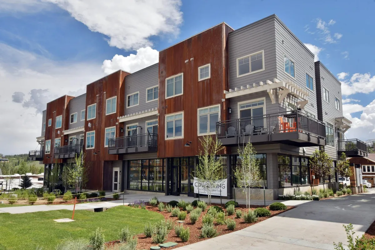 A modern, multi-story building with stylish balconies overlooks a well-designed landscape, bathed in warm sunset light. Visible activity hints at weekly farmers' markets and community festivals, with people gathering and dining outdoors, emphasizing the vibrant local scene and seasonal events available year-round.