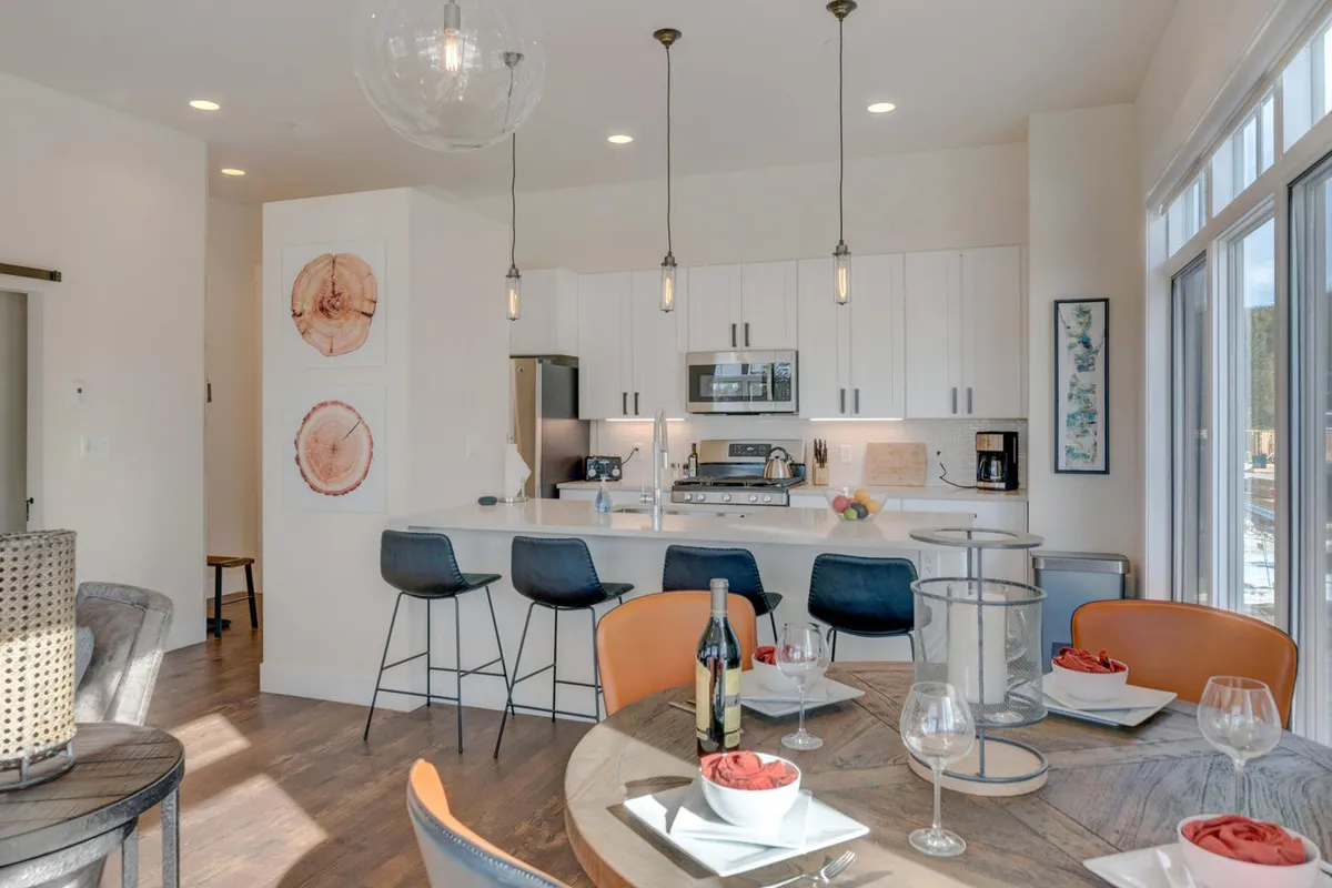A spacious, contemporary kitchen and dining area with a large kitchen island, modern appliances, a sleek dining table set for a meal, vibrant wall art, and expansive windows flooding the space with natural light. The setting is visibly ready for cooking, dining, and entertaining guests, providing a sense of warmth and togetherness.