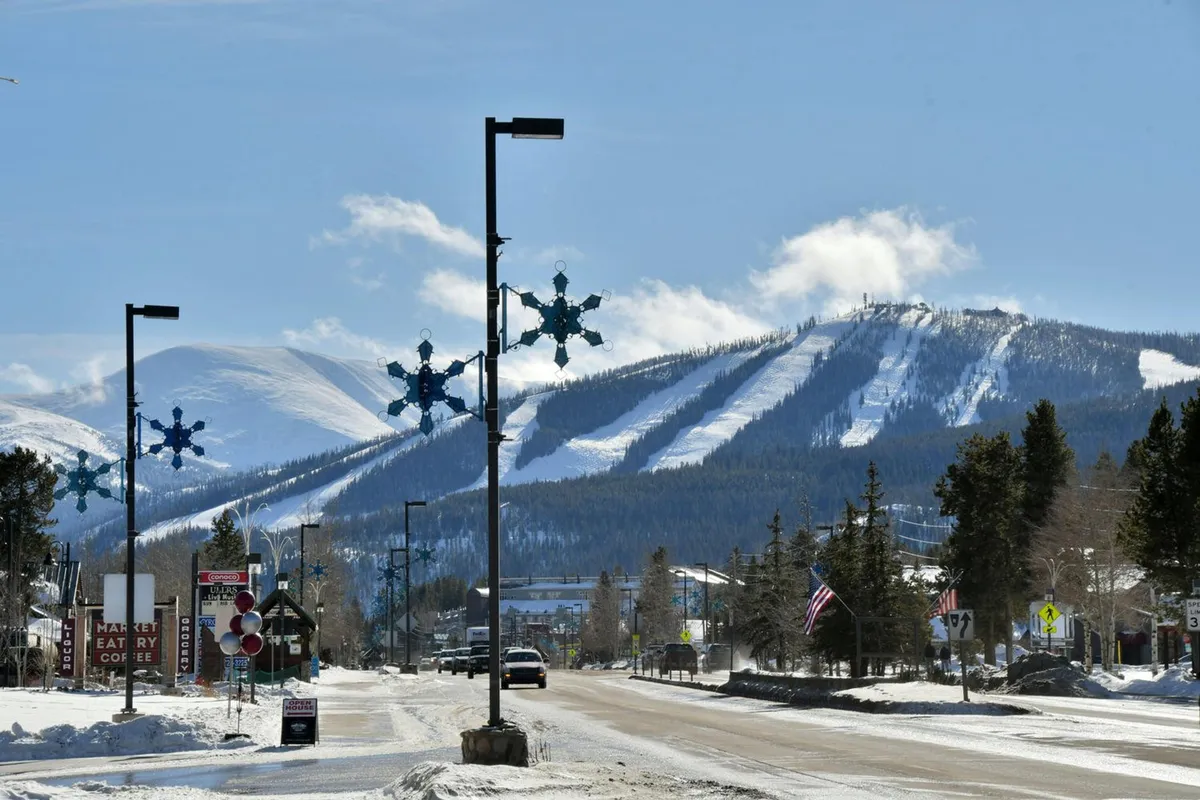 A festive winter scene on the main street, with snow-covered mountains in the background, decorative snowflake lights, welcoming shops, and a lively street ambiance. People are seen enjoying the winter atmosphere, hinting at seasonal activities and the charm of Hideaway Park nearby.